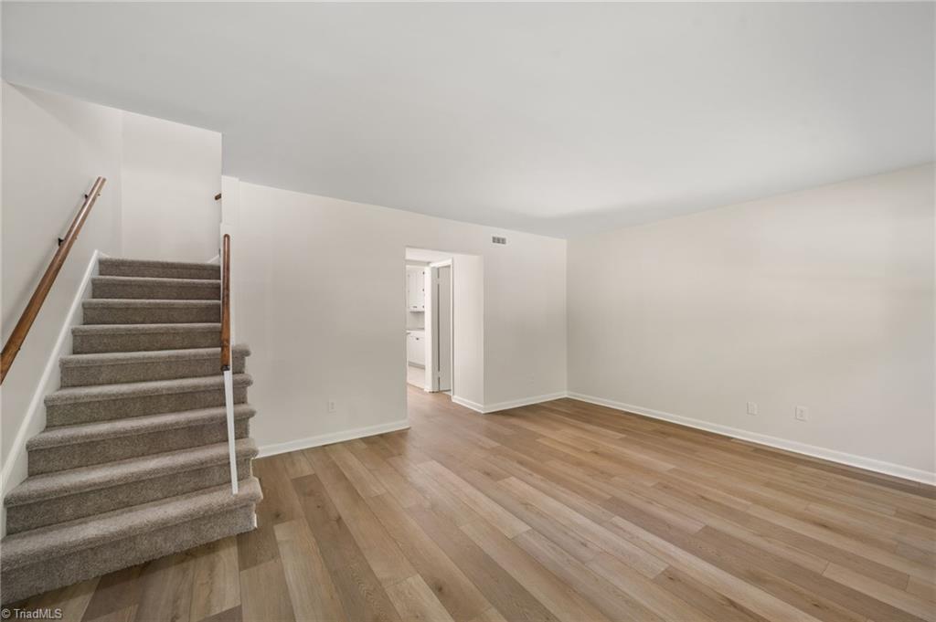 Vacant living room with wood-look flooring, carpeted staircase, and doorway to the kitchen.