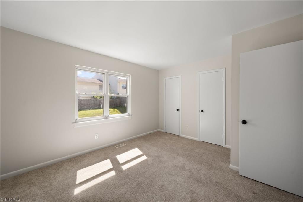 Empty carpeted bedroom with a double window overlooking a fenced lawn and sunlight across the floor.