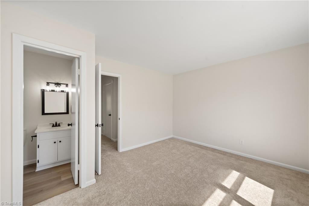 Empty carpeted bedroom with an open doorway to a bathroom vanity and sunlight across the floor.