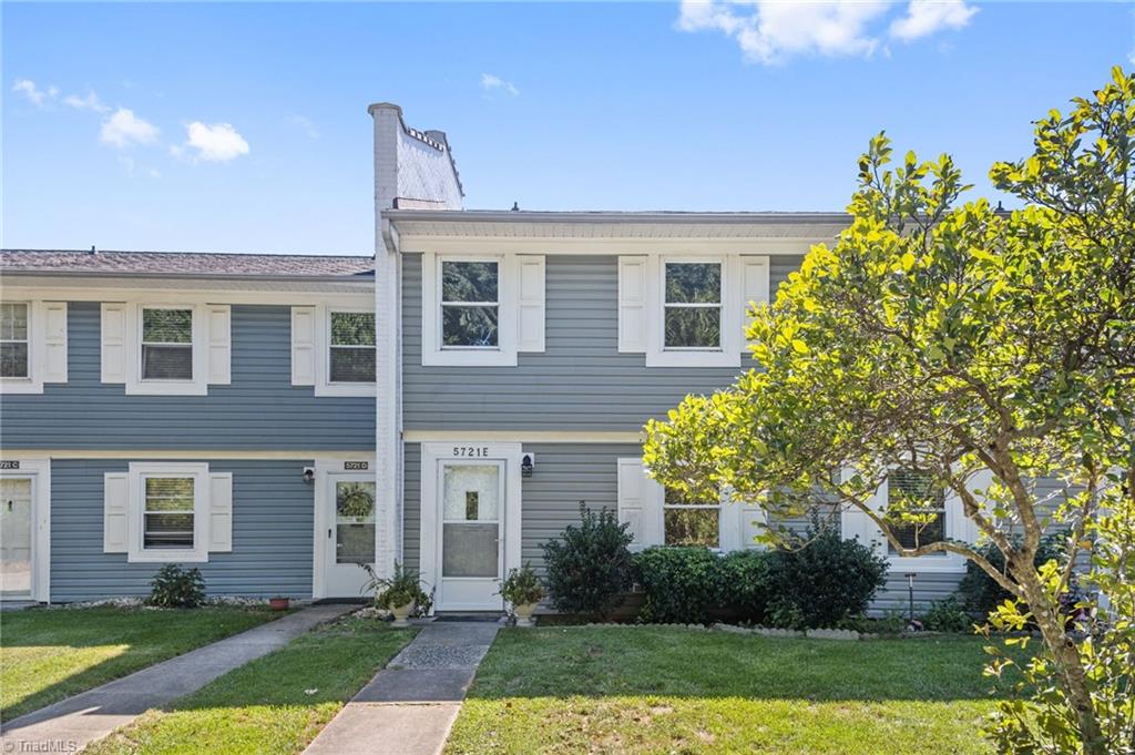 Blue-gray two-story attached home with white shutters, a front walkway, and a leafy tree.