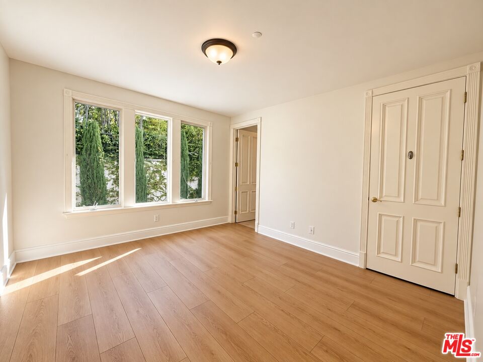 Empty bedroom with light wood floors, three tall windows, and paneled closet doors.