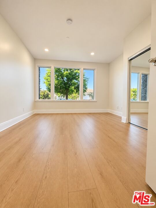 Empty room with light wood floors, broad windows, and leafy trees outside.