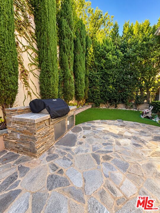 Backyard patio with stone paving, a built-in outdoor kitchen, a small lawn, and tall cypress trees.