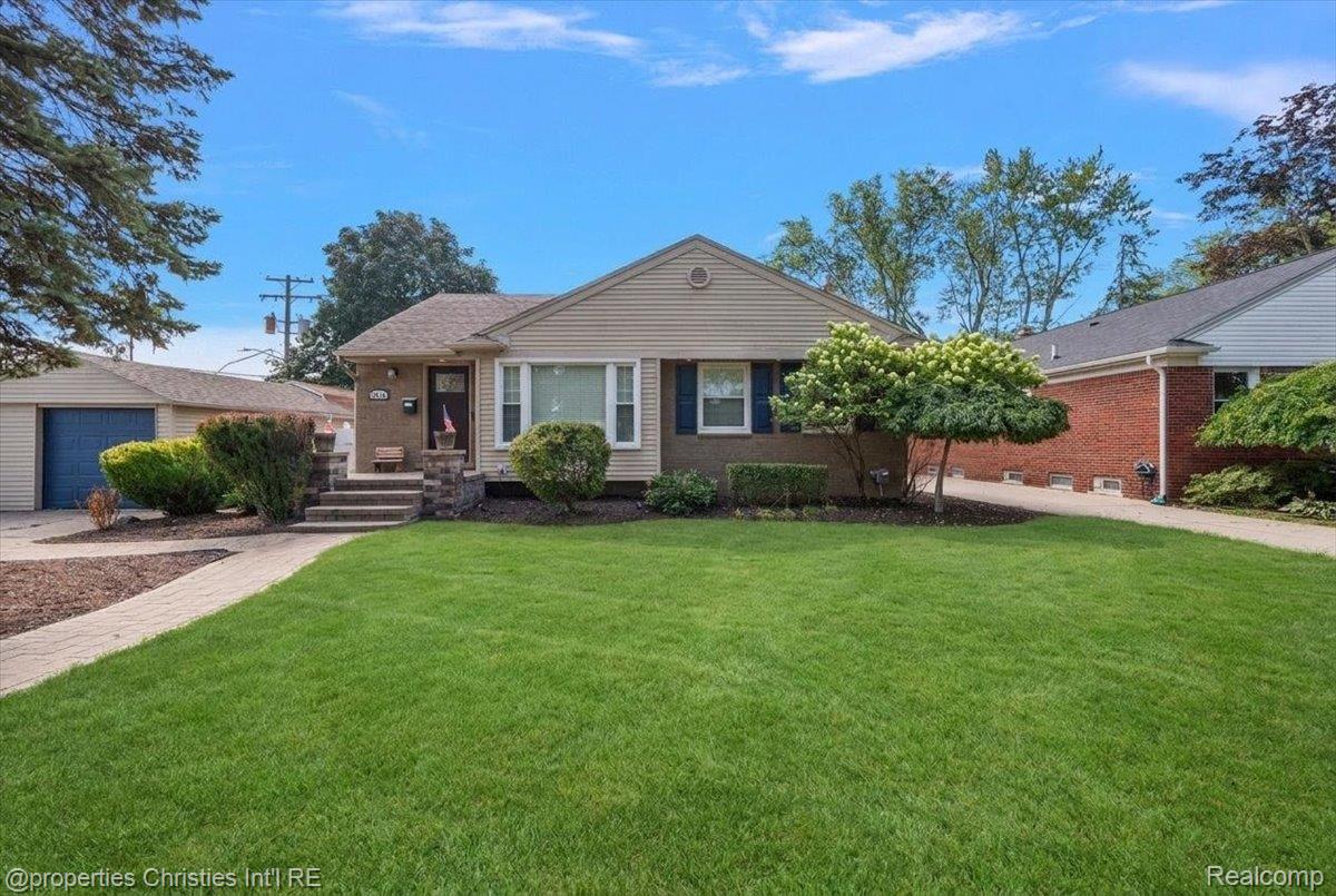 Single-story beige house with blue shutters, landscaped shrubs, and a broad front lawn.