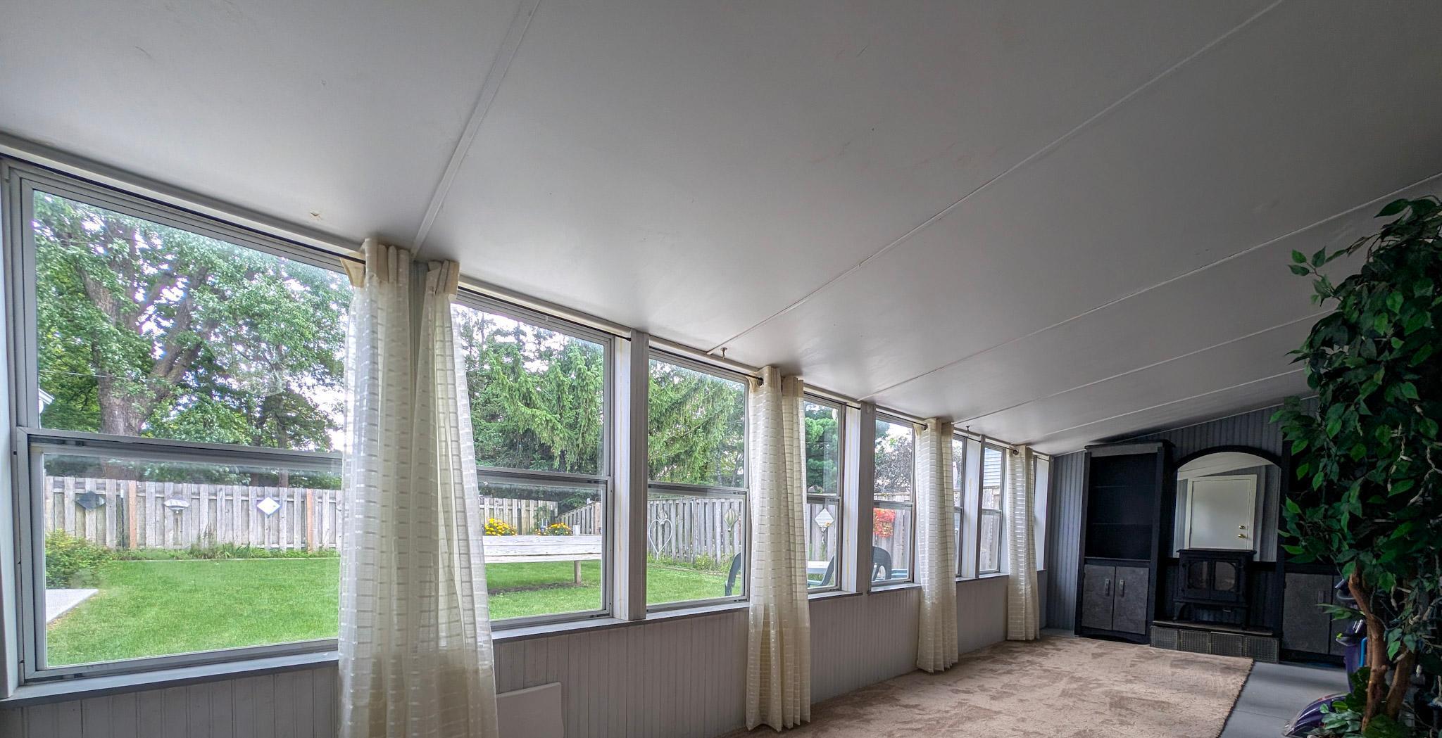 Sunroom with a long row of curtained windows overlooking a fenced yard and a wood stove.