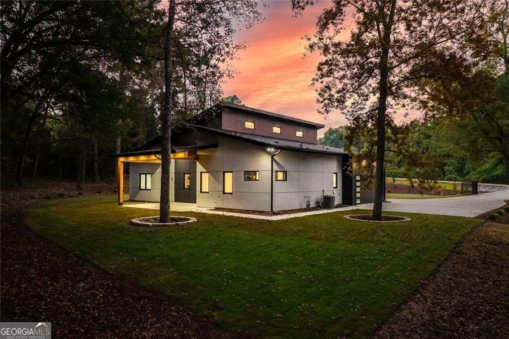 Modern two-story home with a covered entrance and illuminated windows among mature trees at dusk.