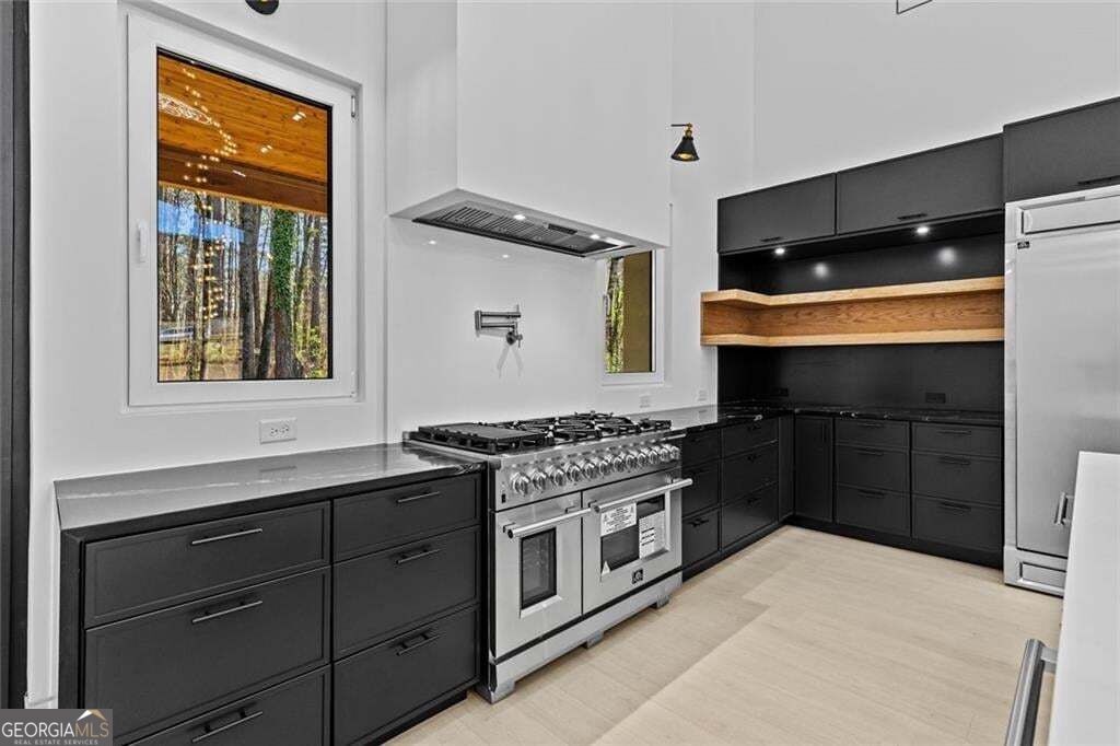 Modern kitchen with dark cabinets, a stainless gas range, wood shelf, and window overlooking trees.
