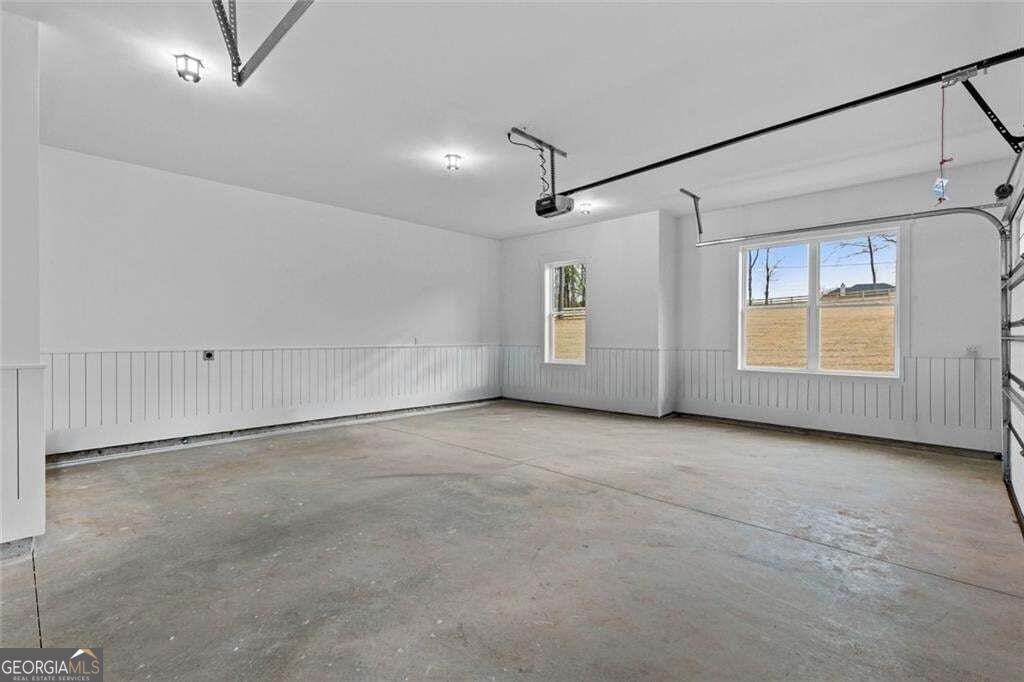 Empty garage with concrete flooring, white paneled walls, and windows overlooking the yard.