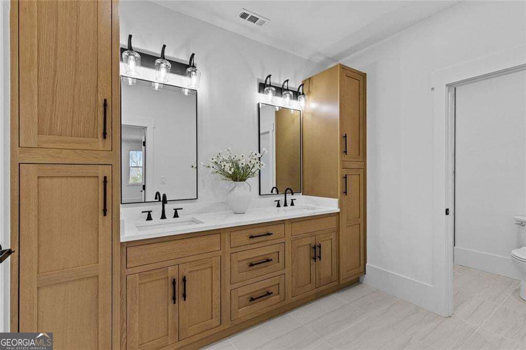 Bathroom with a double vanity, light wood cabinetry, black faucets, and a vase of flowers.
