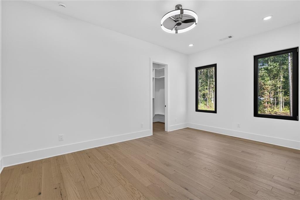 Empty bedroom with light wood floors, ceiling fan, and black-framed windows overlooking trees.