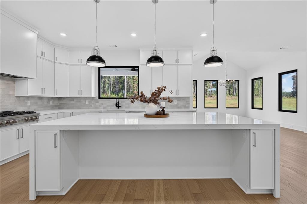Modern white kitchen with a large island, black pendant lights, and windows overlooking trees.