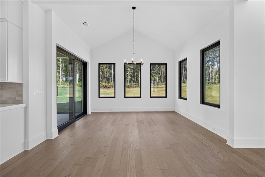 Empty dining area with vaulted ceiling, chandelier, and windows overlooking a grassy yard and trees.