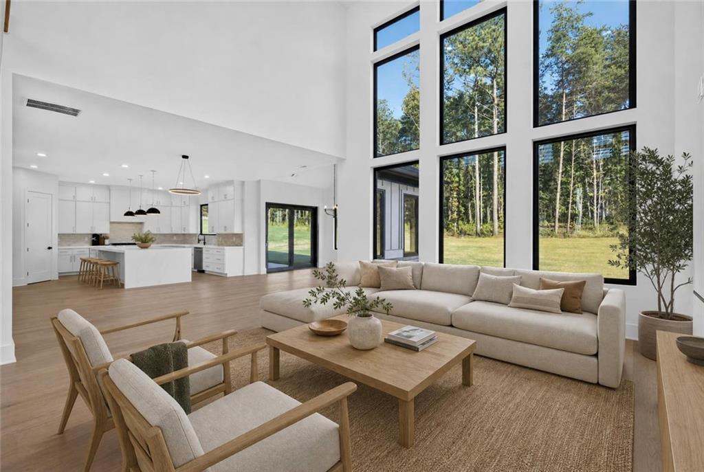 Staged living room with soaring windows, neutral seating, and an open kitchen overlooking trees.