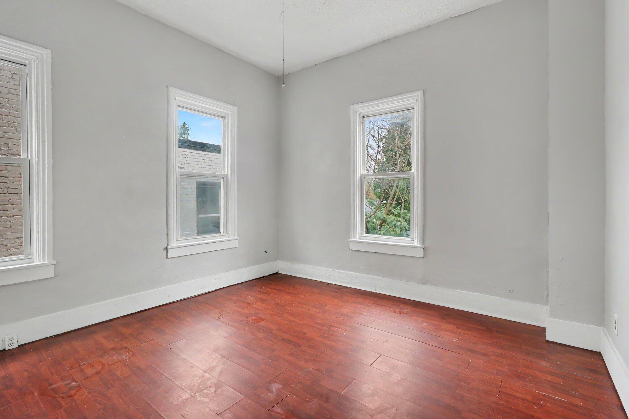 Empty room with reddish-brown hardwood floors, gray walls, and three tall windows.