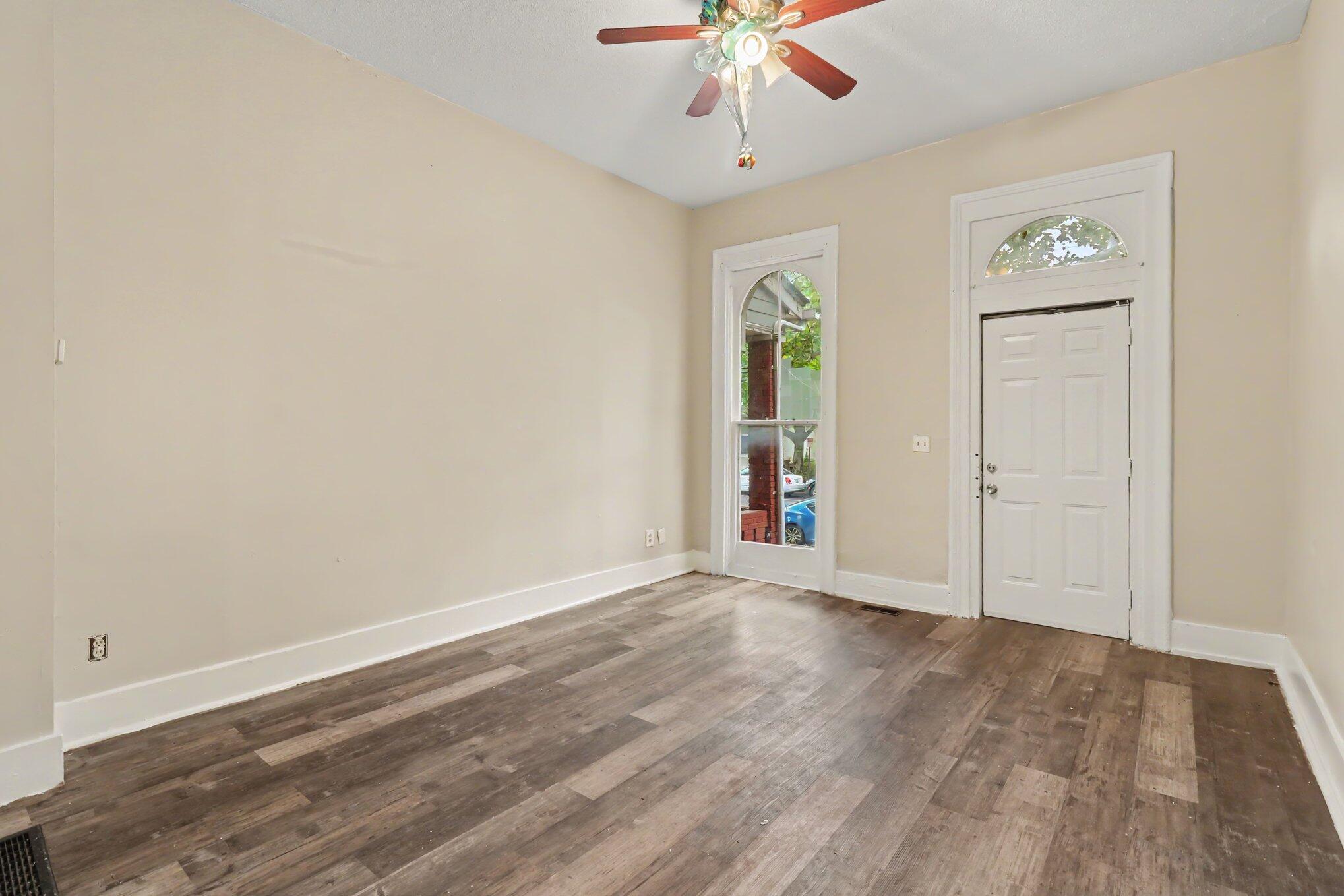 Empty front room with dark plank flooring, a ceiling fan, and a white entry door beside an arched window.