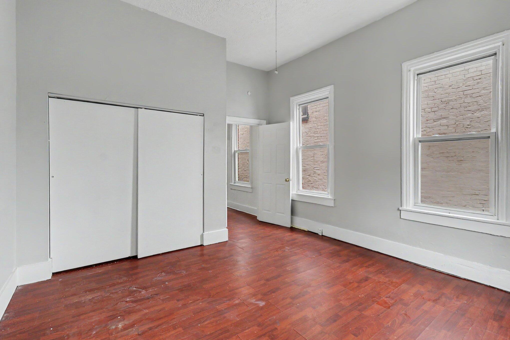 Empty room with reddish hardwood floors, sliding closet doors, and tall windows.