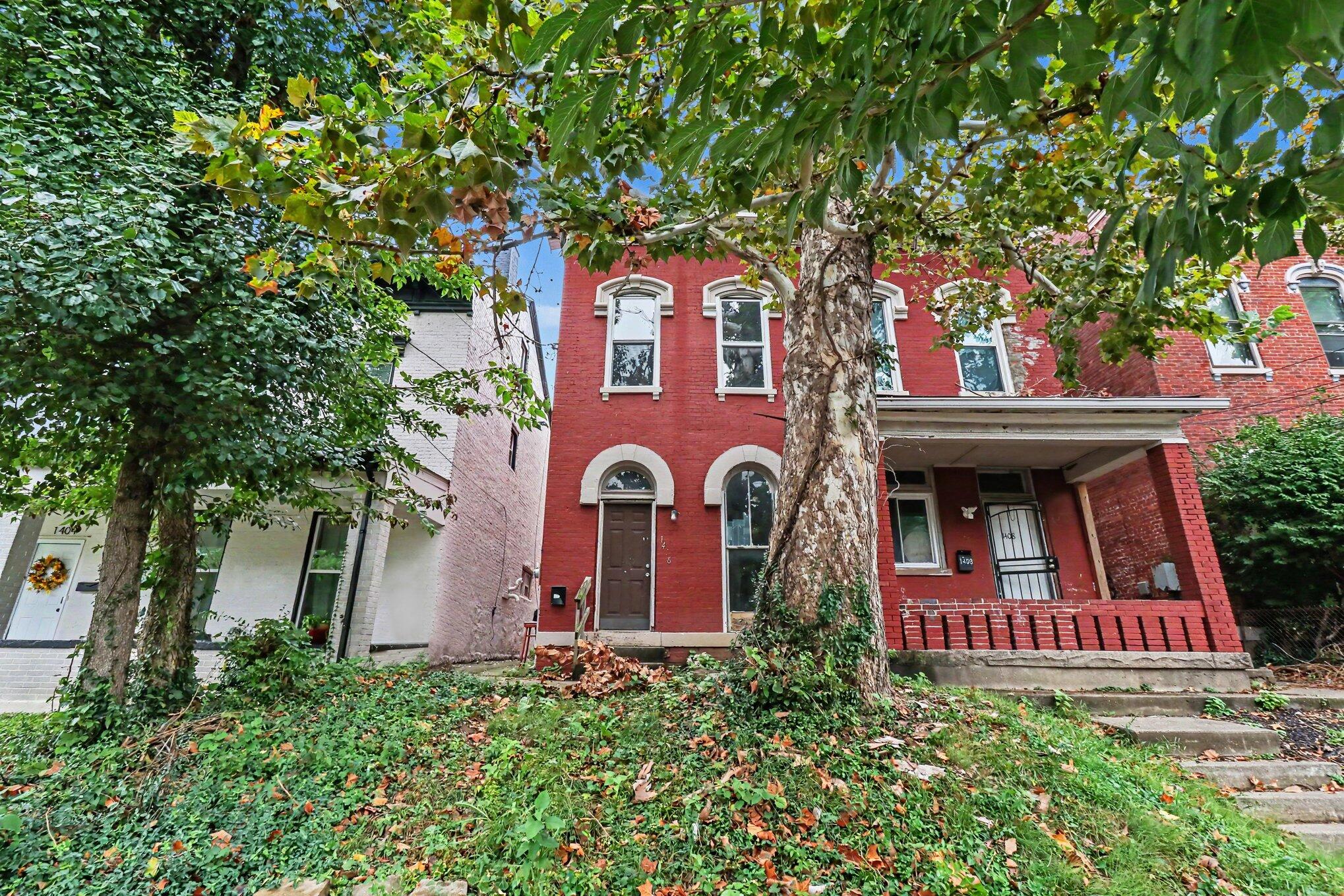 Red brick home with arched windows and a covered porch, partly obscured by a large tree.
