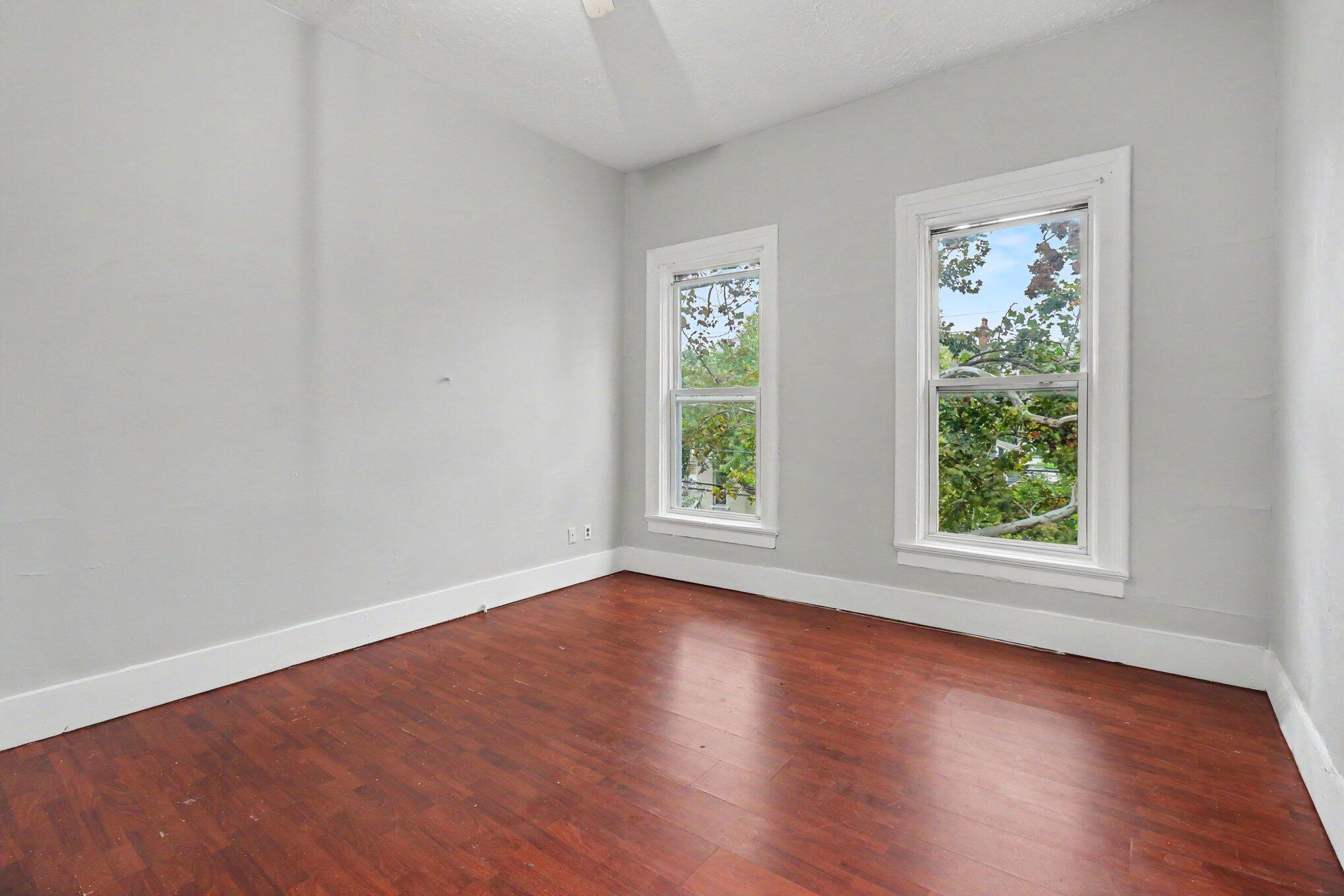 Empty room with reddish-brown hardwood floors and two tall windows.