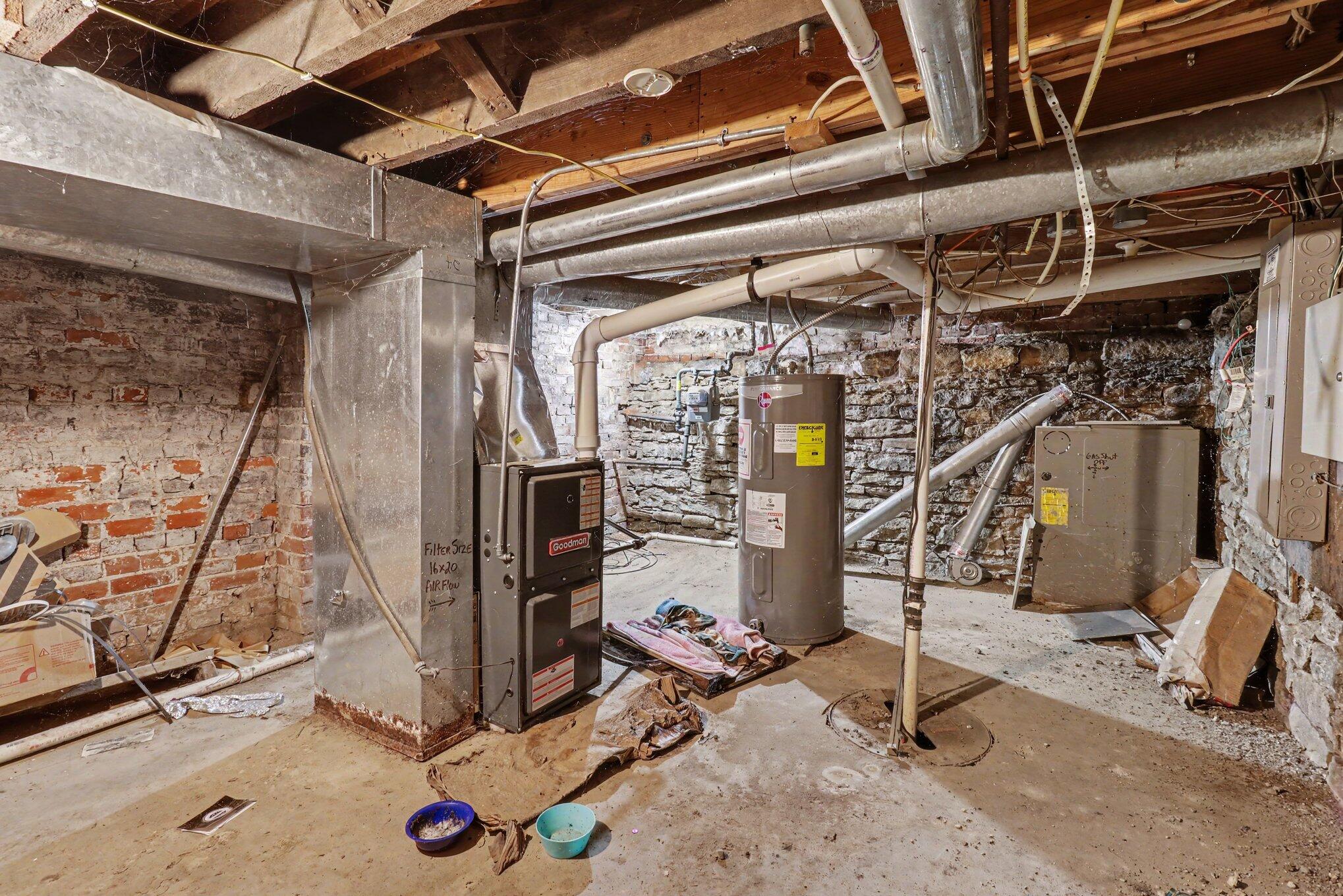 Basement mechanical room with exposed brick and stone walls, overhead ductwork, furnace, and water heater.