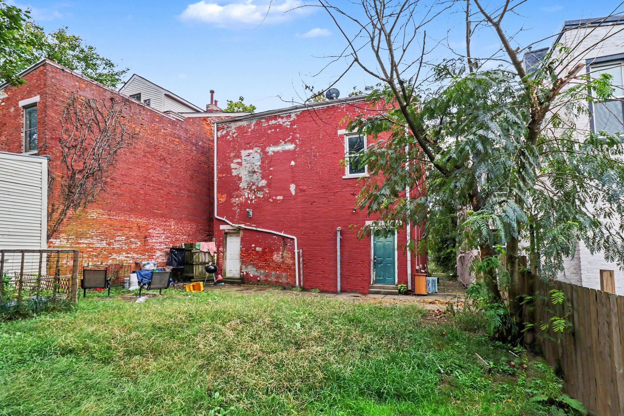 Red brick rear exterior with peeling paint, a teal door, and a grassy, tree-shaded backyard.