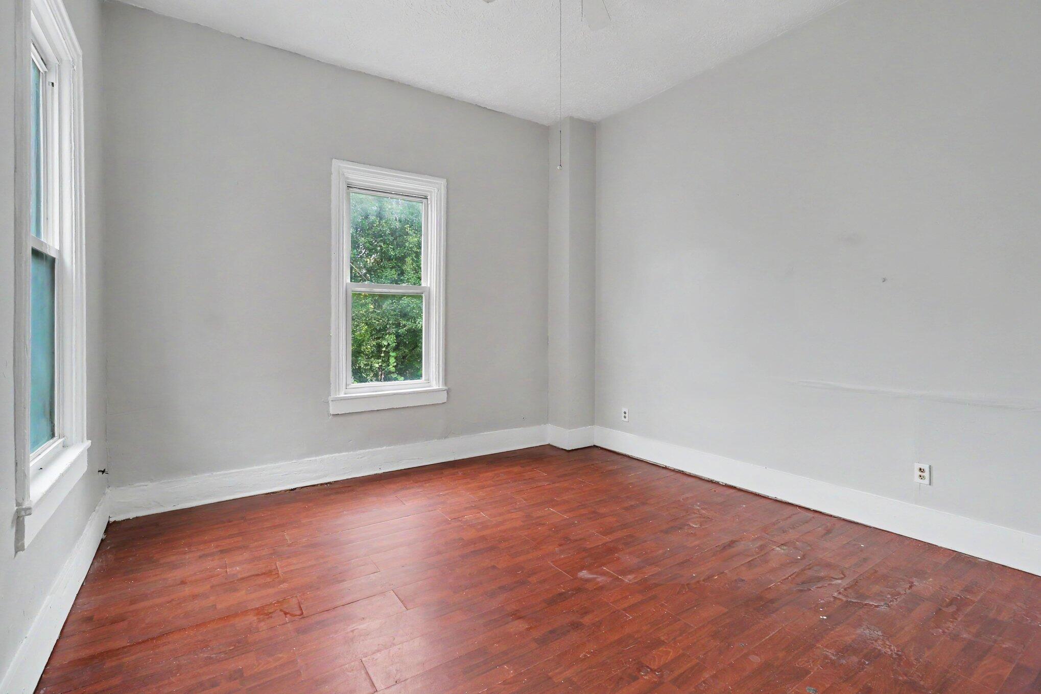 Empty room with gray walls, two windows, and reddish-brown hardwood floors.