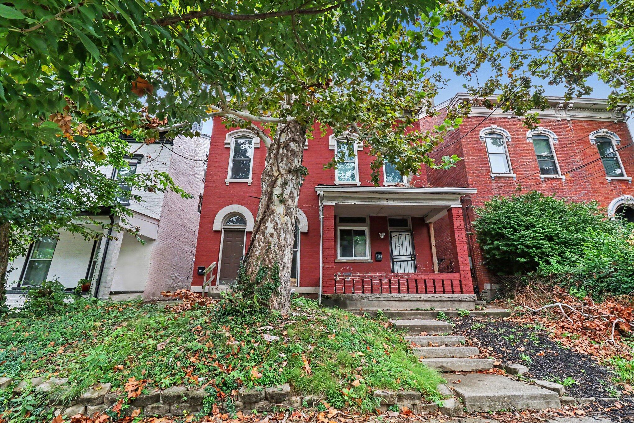 Red-brick two-story home with a covered porch, partly obscured by a mature tree.