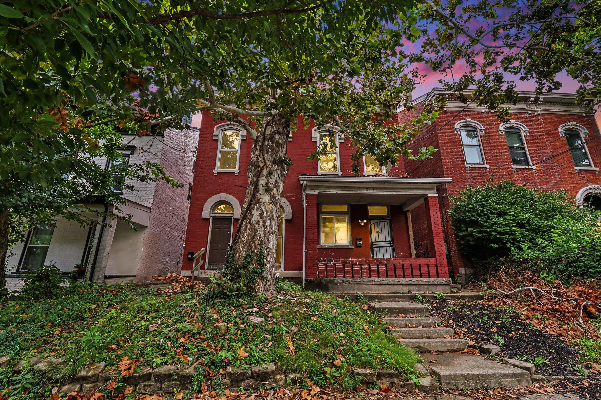 Red brick home with a covered porch and stone steps, framed by mature trees and autumn leaves.