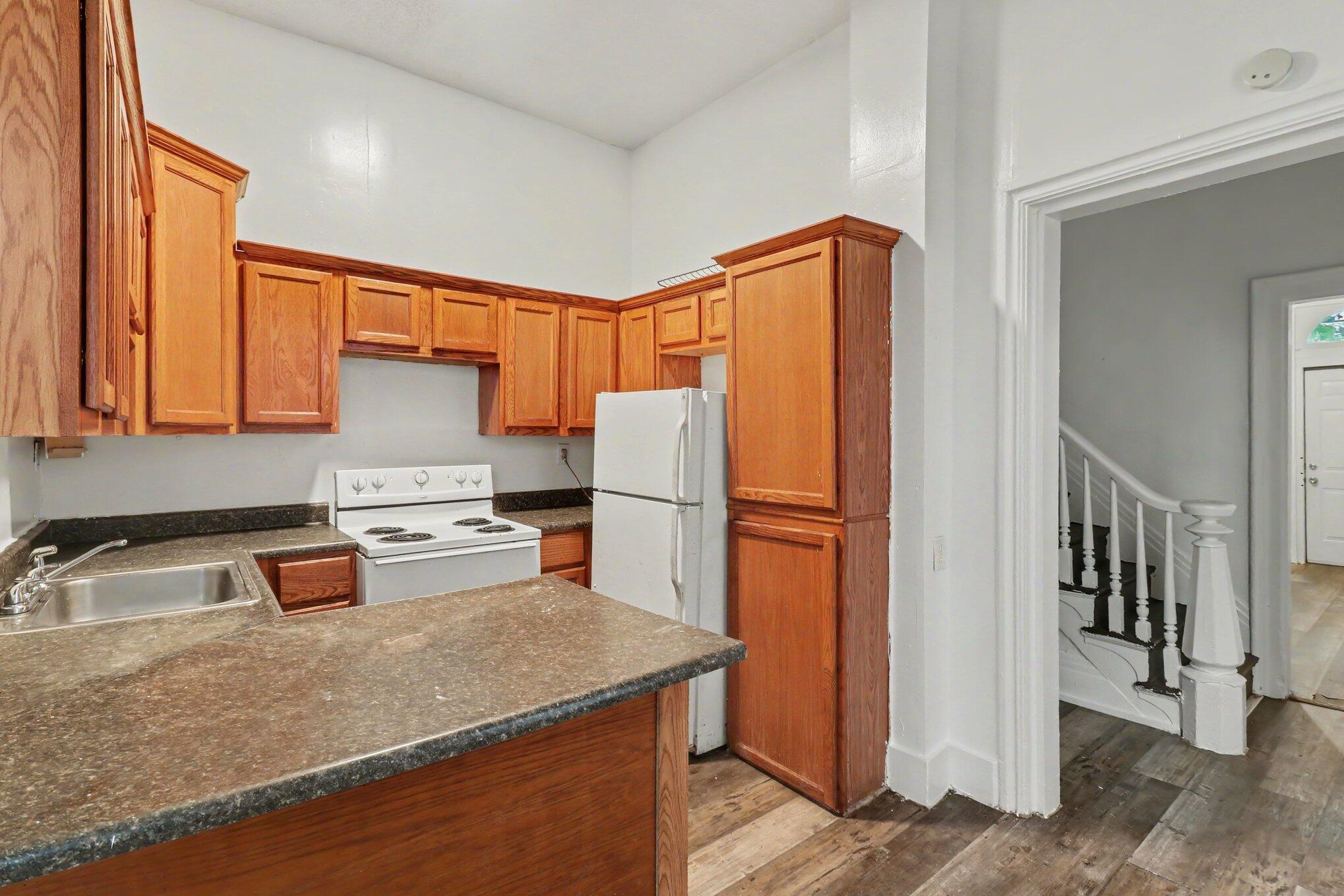 Vacant kitchen with reddish wood cabinets, dark countertops, white appliances, and a doorway to the staircase.