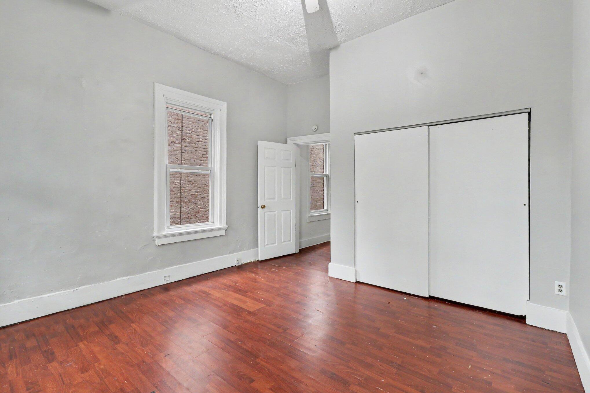 Empty room with hardwood floors, a large sliding-door closet, and windows facing a brick wall.