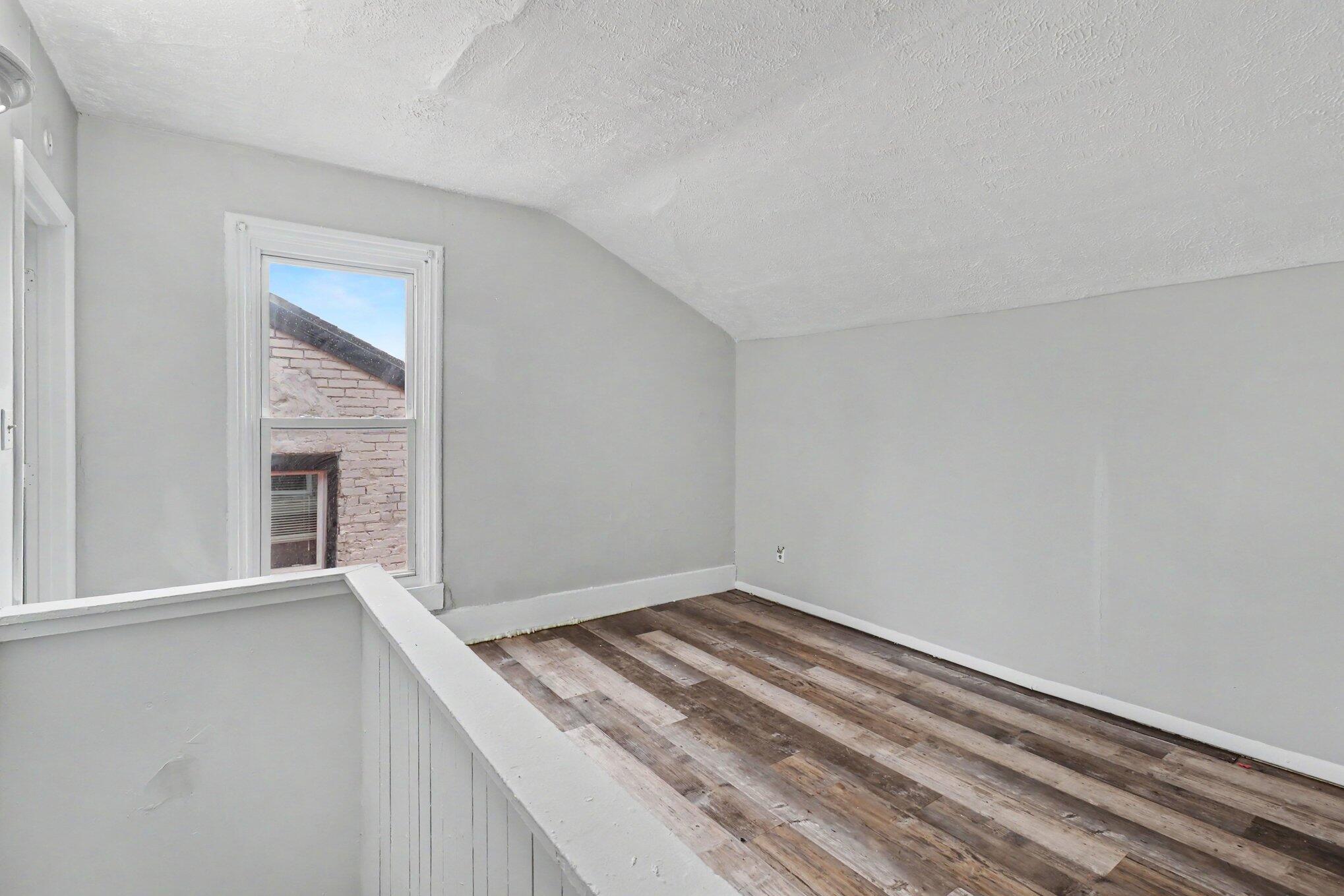 Vacant upstairs room with weathered plank flooring, a sloped ceiling, and a window beside a white half-wall.