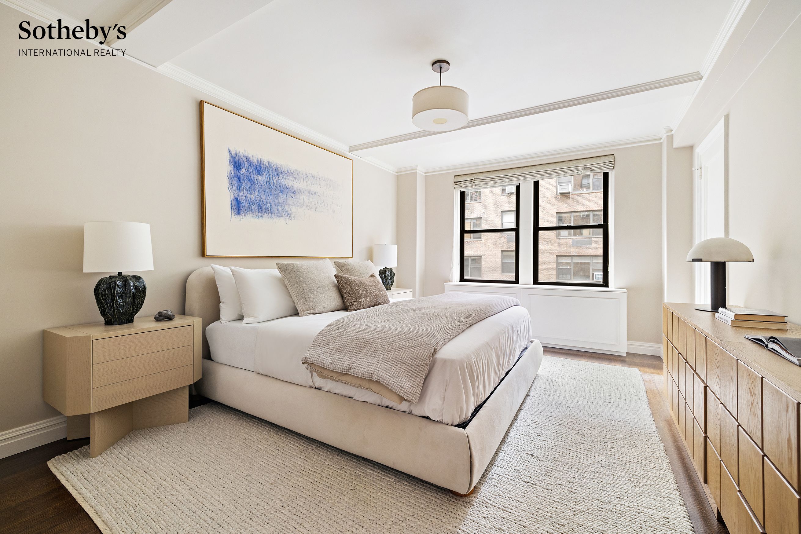 Bright neutral bedroom with a low upholstered bed, blue abstract artwork, and black-framed windows.