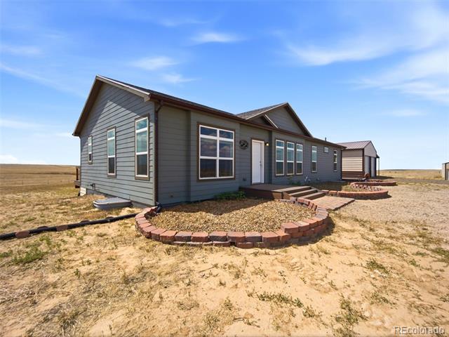 Gray-sided single-story home with a white entry door, brick-edged garden beds, and open prairie surroundings.