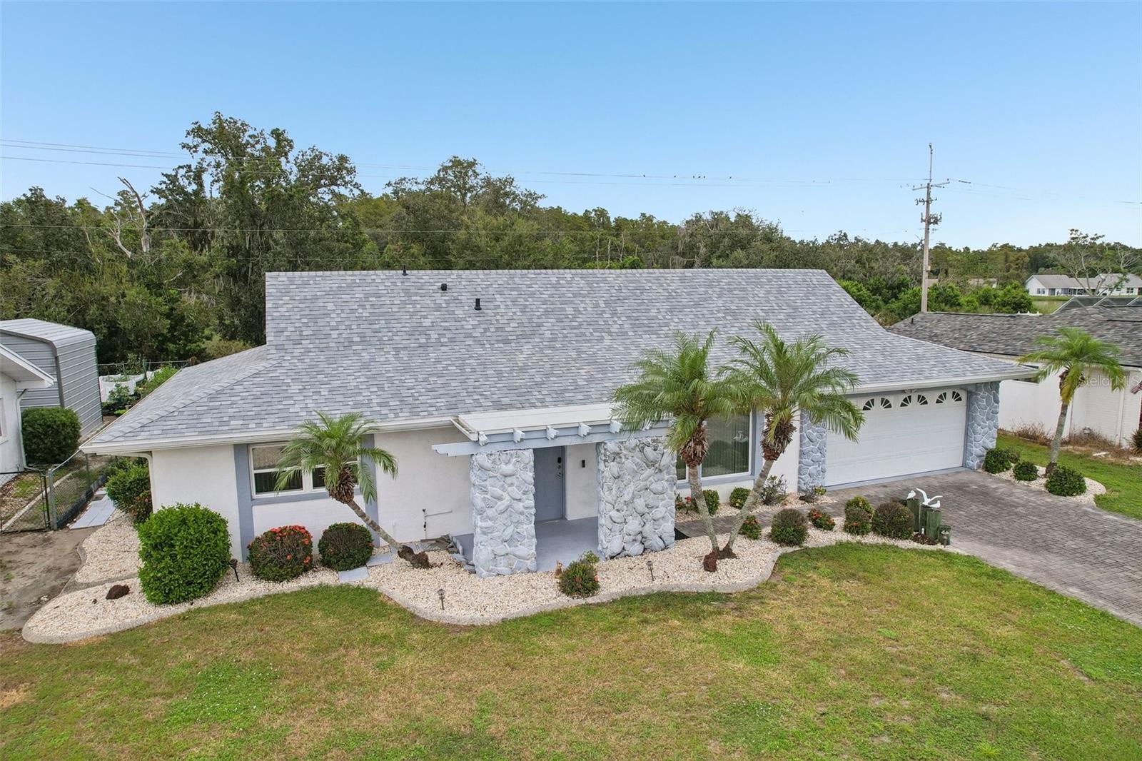 Single-story home with stone entry columns, gray shingle roof, white garage door, and palm trees.
