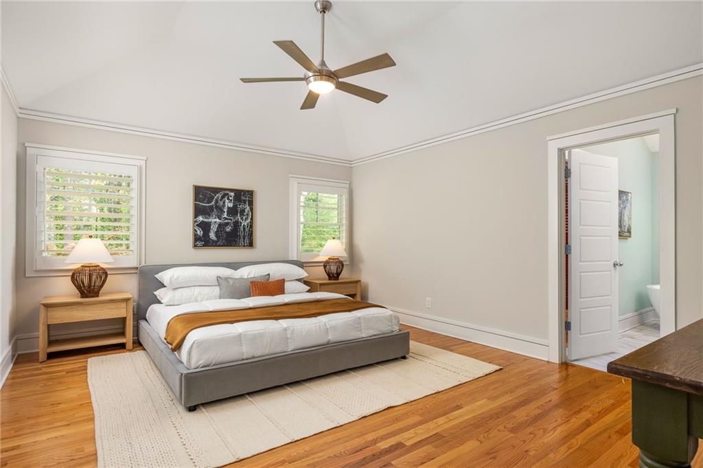 Staged bedroom with a gray bed, white bedding, hardwood floors, and two shuttered windows.
