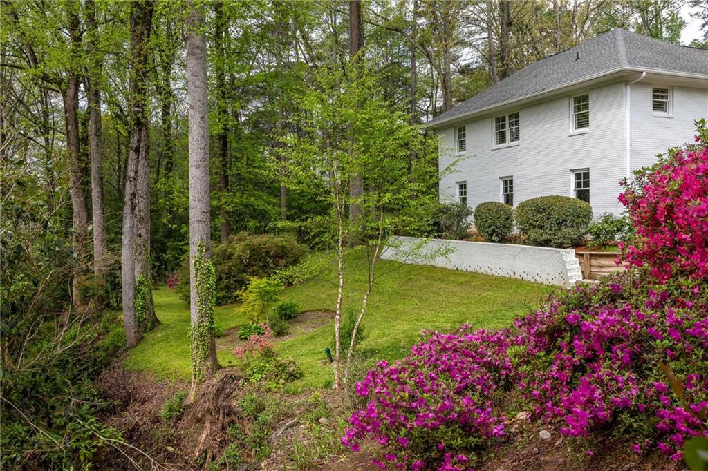 White two-story house on a wooded slope, framed by blooming pink azaleas and a low retaining wall.