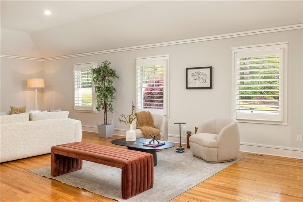 Staged living room with hardwood floors, pale seating, a rust-colored bench, and three shuttered windows.