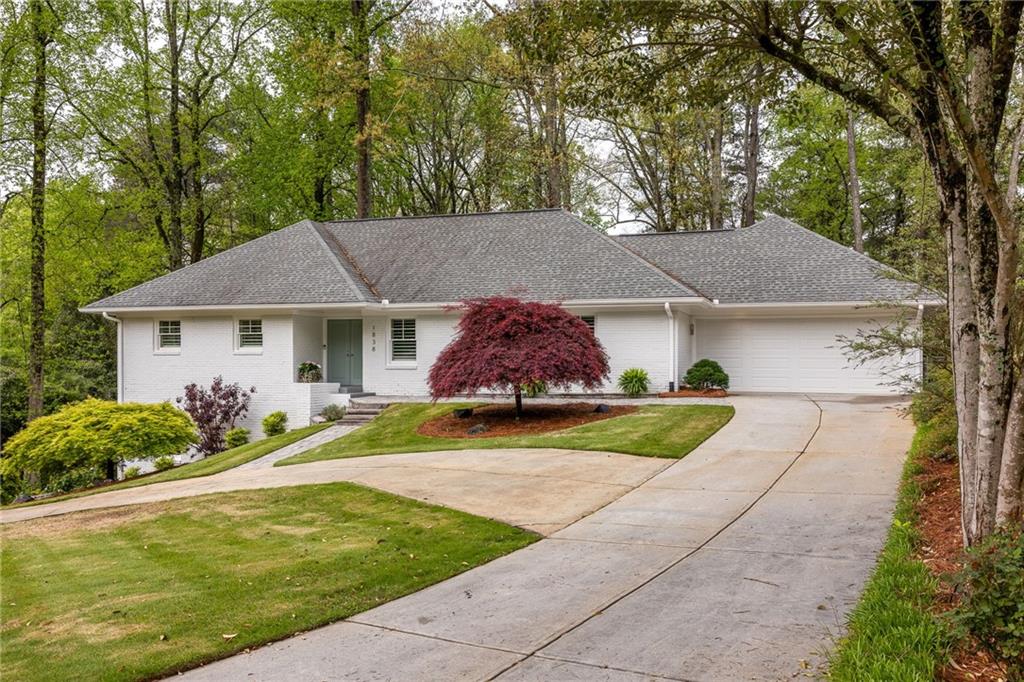 White brick ranch home with a gray roof, attached garage, curved driveway, and red-leaved ornamental tree.