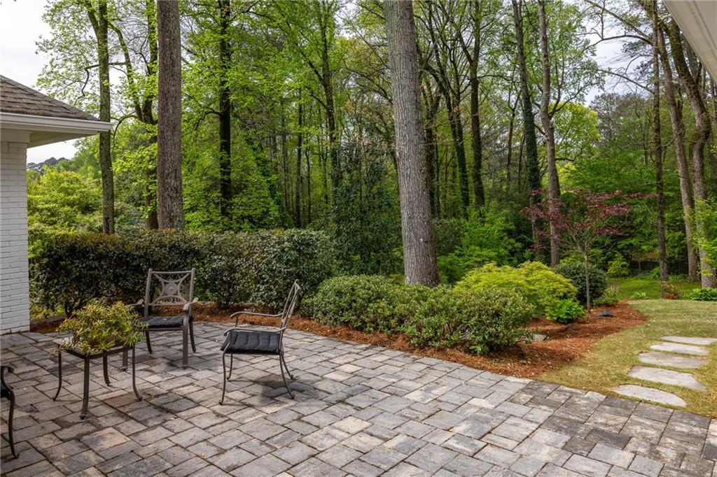 Stone patio with metal chairs, landscaped shrubs, and a wooded backyard.