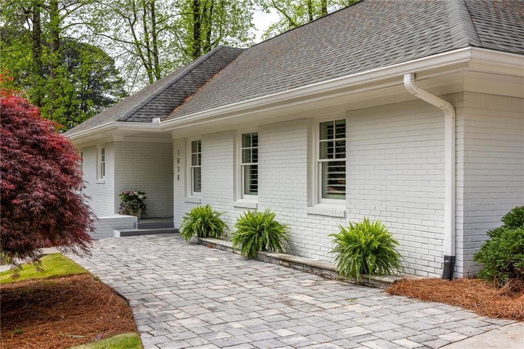 White brick house with a gray shingle roof, stone-paver driveway, and landscaped front entry.
