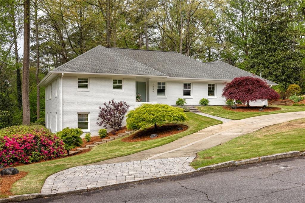 White brick home with a broad gray roof, flowering shrubs, and a sloped front lawn.