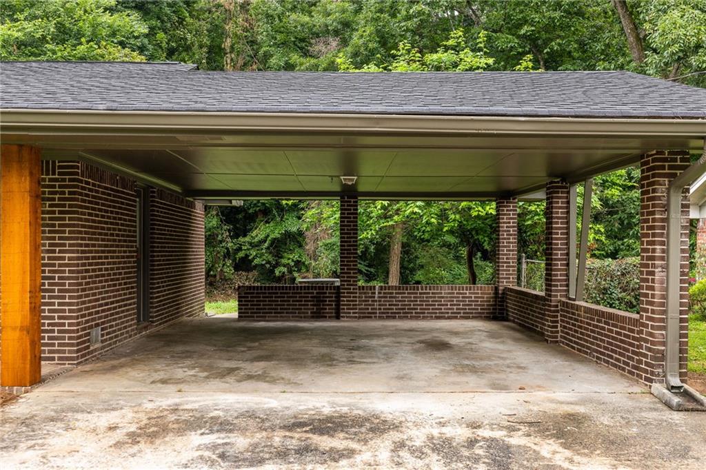 Covered brick carport with a concrete floor and wooded surroundings.