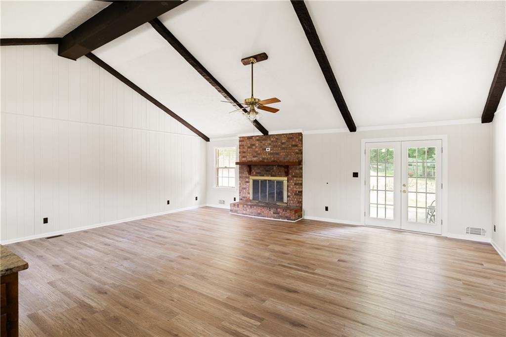 Empty living room with a brick fireplace, vaulted beamed ceiling, wood flooring, and French doors.