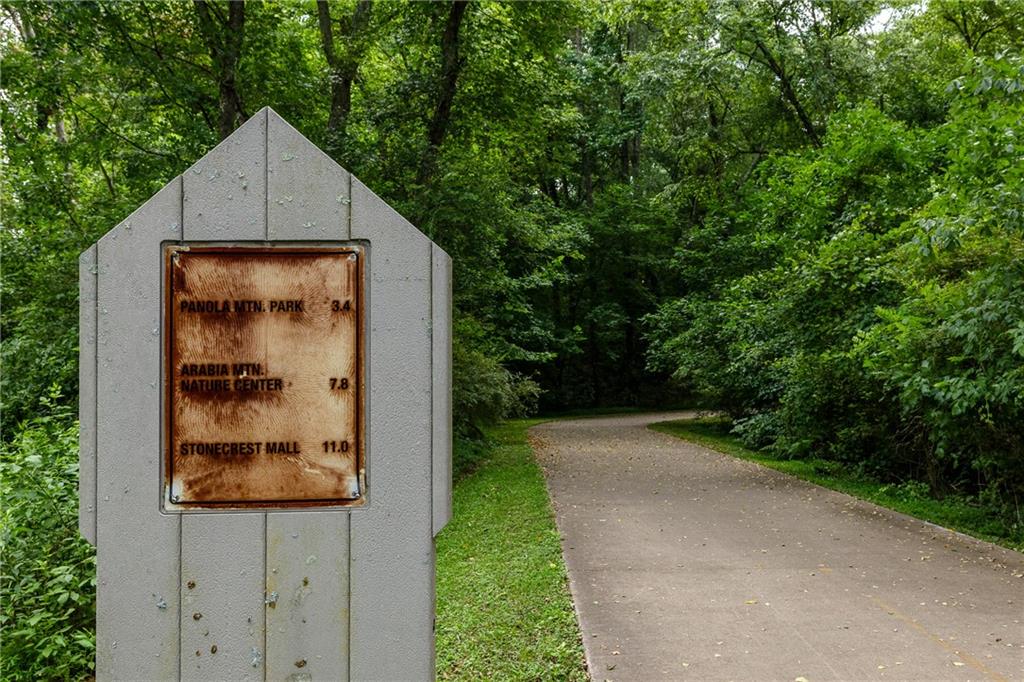 A paved trail curves through dense woods beside a sign showing distances to nearby parks and Stonecrest Mall.
