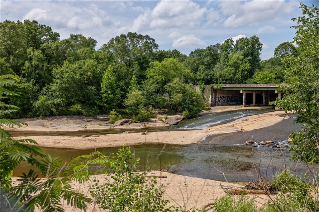 Shallow creek flowing over sandy rock beneath a graffiti-marked bridge, surrounded by dense trees.