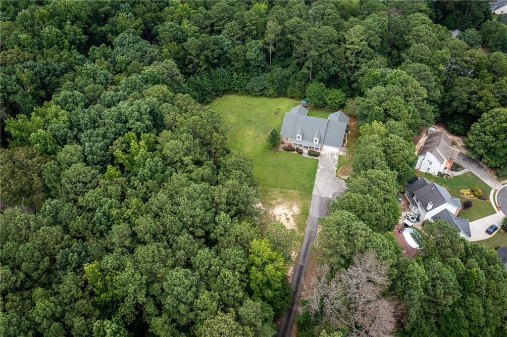 Aerial view of a house on a grassy lot surrounded by dense woods, with a long driveway.