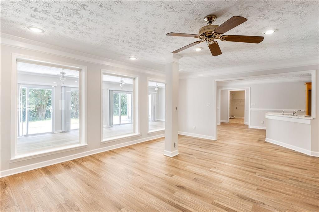Empty living area with light wood floors, ceiling fan, and large windows.