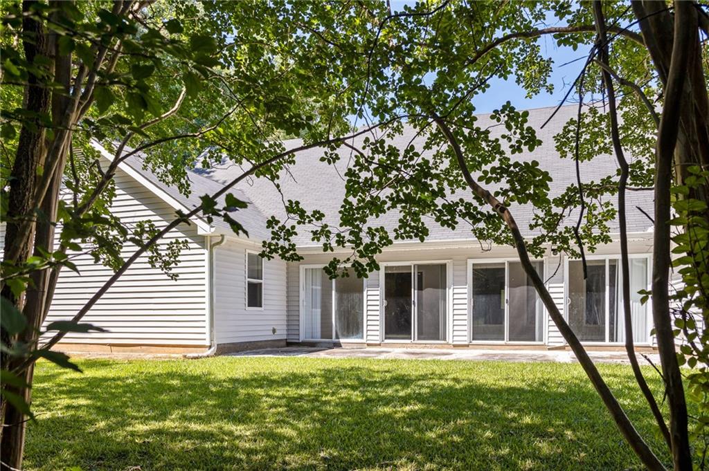 Rear of a white-sided home with sliding glass doors, a patio, and a tree-shaded lawn.