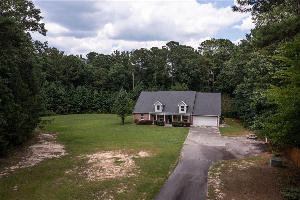 Aerial view of a brick house with front dormers, an attached garage, and a long driveway on a wooded lot.