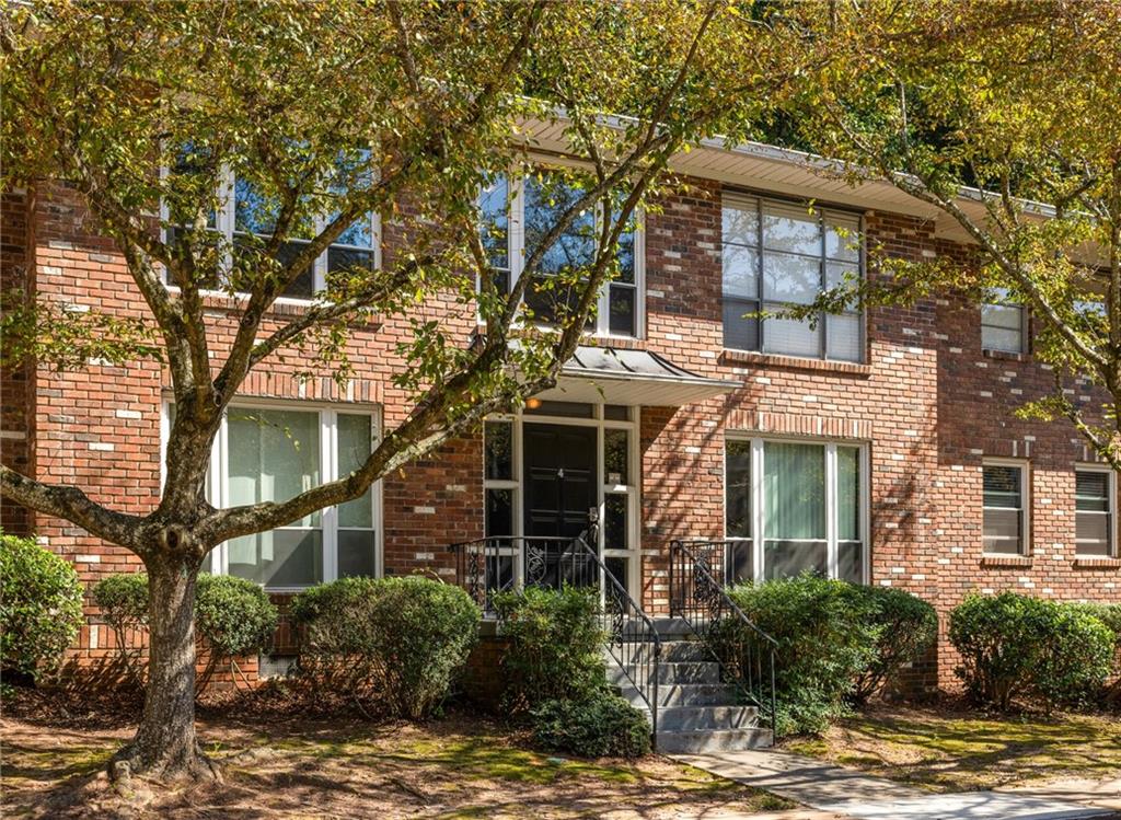 Brick condominium entrance with steps and railings, framed by mature trees and trimmed shrubs.