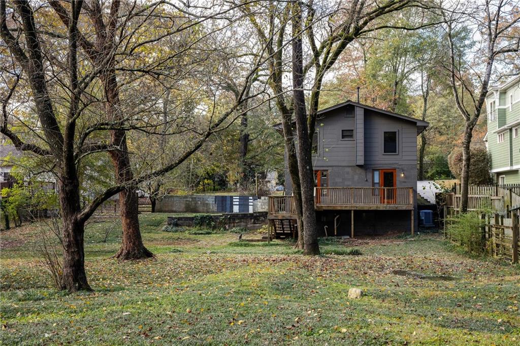 Gray two-story house with a wooden deck in a spacious yard beneath mature trees.